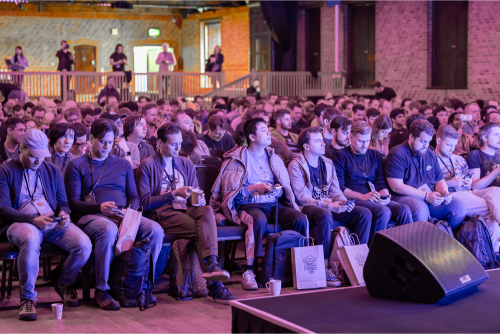 A packed audience seated under purple lighting, watching a talk.