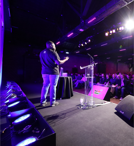 A speaker on stage seen from the wings, addressing the room under stage lights.